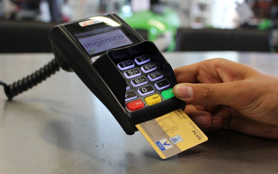 Mano de una persona de tez morena pagando en una terminal color negra con botones de colos rojo, amarillo y verde con una tarjeta de color oro para ejemplicar de cuanto es la multa por comisión de pago con tarjeta.